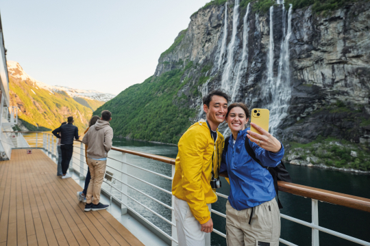 Ein Paar steht an einer Reling und macht ein Selfi von sich vor den Bergen im Hintergund 